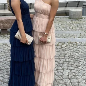 Beige oneshoulder klänning - Elegant beige rosa oneshoulder klänning med volanger. Perfekt för en festlig kväll. Klänningen har en lång design och är tillverkad i ett lätt och luftigt material.