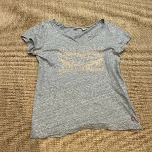 Grå t-shirt från Levi's - Säljer en grå t-shirt från Levi's i bomull. Den har en v-ringad hals och ett tryck på framsidan. Perfekt för en avslappnad stil. T-shirten är i bra skick och passar till vardags.