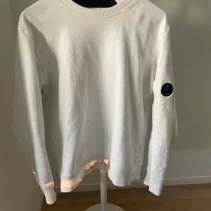 Vit tröja från C.P. Company - Säljer en stilren vit tröja från C.P. Company i mycket bra skick. Tröjan har en rund halsringning och en cool detalj med en svart patch på ärmen. Perfekt för både vardag och lite mer avslappnade tillfällen. Passar bra till jeans eller chinos!