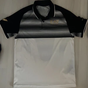 Svartvit randig pikétröja från EA7 (slutsåld överallt!) - Säljer en  pikétröja från EA7 i svart och vitt med randigt mönster upptill. Tröjan har en klassisk krage och korta ärmar, perfekt för både vardag och sportiga aktiviteter. Jätte eftertraktad tröja som är slutsåld överallt online! Den är i mycket bra skick och har en stilren design med EA7-loggan på bröstet och på armen. 