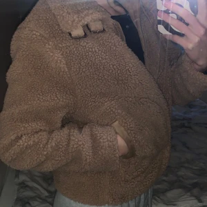 Beige teddyjacka med dragkedja - Mysig beige teddyjacka i mycket bra skick. Den har en dragkedja framtill och dekorativa spännen vid kragen. Jackan har långa ärmar och en skön, varm känsla. 