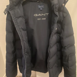 Svart dunjacka från GANT - Säljer en snygg och varm svart dunjacka från GANT. Jackan har en huva och dragkedja framtill. Perfekt för kalla vinterdagar. Den är i mycket bra skick och har en stilren design med GANT-loggan på bröstet och ryggen. Storlek 170 men passar även för de som har storlek S.