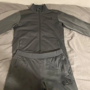 Grå träningsdräkt från EA7 Emporio Armani - Säljer en grå träningsdräkt från EA7 Emporio Armani. Setet består av en långärmad tröja med dragkedja och matchande byxor.