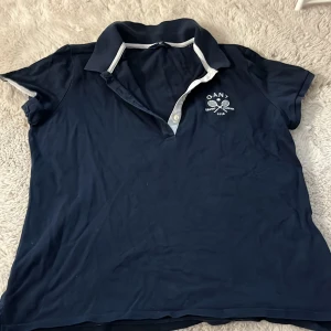 Mörkblå pikétröja från Gant - Säljer en snygg mörkblå pikétröja från Gant med vit broderad logga och tennismotiv på bröstet. Tröjan har en klassisk krage med vita detaljer och korta ärmar. Perfekt för en avslappnad men stilren look!