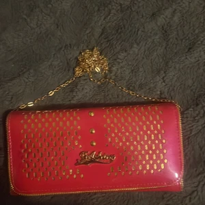 Rosa plånbok med guldkedja - (PRISET KAN SÄNKAS) Snygg rosa plånbok med guldfärgade detaljer och kedja. Den har flera fack för kort och en dragkedja för mynt. Perfekt för att hålla ordning på dina saker med stil. Den glänsande ytan och gulddetaljerna ger en lyxig känsla. Bred-20cm längd-10cm