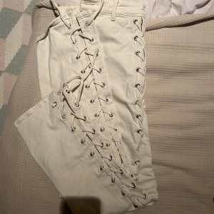 Beige byxor med snörning - Snygga beige byxor med unik snörning längs sidorna. Byxorna har en hög midja och knappar framtill. Perfekta för en trendig look.