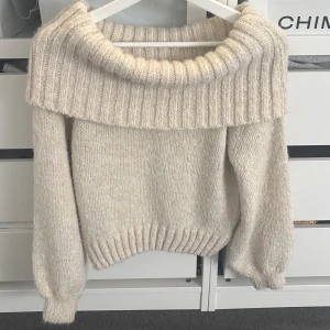 Beige stickad offshoulder tröja - Superfin beige stickad tröja med offshoulder-design. Perfekt för höst och vinter, den är både mysig och stilren. Tröjan har långa ärmar och en bred ribbad kant som ger en extra touch. Passar perfekt till både jeans och kjol för en avslappnad men chic look.