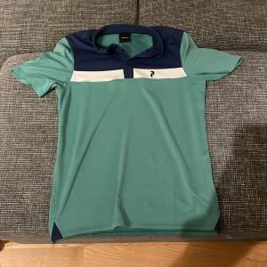 Säljer en snygg pikétröja från Peak Performance i grönt och marinblått. Tröjan har en klassisk krage och korta ärmar, perfekt för både vardag och sport. Den är i mycket bra skick och har en stilren design med en vit rand över bröstet.Pris går alltid att förhandlas!