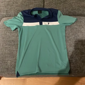Grön och marinblå pikétröja från Peak Performance - Säljer en snygg pikétröja från Peak Performance i grönt och marinblått. Tröjan har en klassisk krage och korta ärmar, perfekt för både vardag och sport. Den är i mycket bra skick och har en stilren design med en vit rand över bröstet.Pris går alltid att förhandlas!