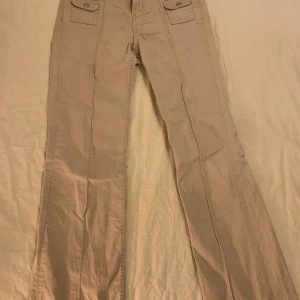 Beige byxor med fickdetaljer - Säljer ett par snygga beige byxor i bra skick. De har en bootcut-stil och praktiska fickor med knappar framtill. Perfekta för både vardag och festliga tillfällen. Passar bra till höst- och vårgarderoben!