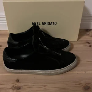 Svarta sneakers från Axel Arigato - Snygga svarta sneakers från Axel Arigato med dragkedja framtill. Perfekta för en stilren look. Skorna har en vit sula som ger en fin kontrast till det svarta.