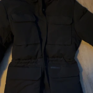 Svart dunjacka - Säljer en svart dunjacka i mycket bra skick. Den har långa ärmar och praktiska fickor framtill. Perfekt för kalla vinterdagar. Jackan har en stilren design med dragkedja och knappar för extra skydd mot kylan. Har använt fåtal gånger storlek S