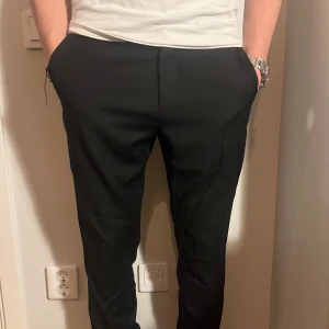 Svarta kostymbyxor från Whyred - Snygga svarta kostymbyxor från Whyred i storlek 44. Perfekta för både formella och semi-formella tillfällen. Byxorna har en klassisk passform med knappar och dragkedja. Tillverkade i Portugal, vilket garanterar hög kvalitet. Ett måste i garderoben för en stilren look!