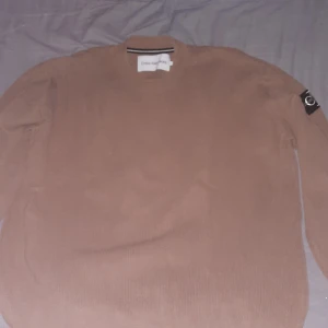 Beige tröja från Calvin Klein Jeans - Säljer en snygg beige tröja från Calvin Klein Jeans i storlek L. Tröjan är ribbad och har en stilren design med en CK-logga på ärmen. Perfekt för både höst och vår, och passar till många olika outfits. Skön och enkel att bära!
