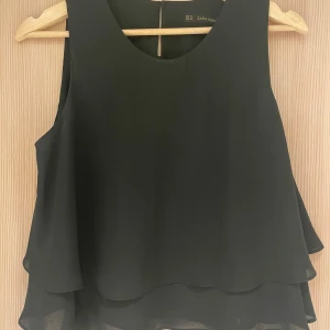 Svart topp från Zara - Snygg och svart ärmlös volang topp från Zara. Säljer då den tyvärr inte kommer till användning. Köparen står för frakt.