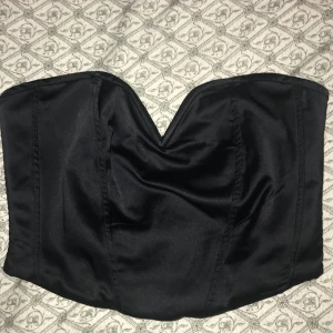 Svart tubtopp från Gina Tricot - Snygg svart tubtopp från Gina Tricot i ett glansigt material. Toppen har en dragkedja bak och en stilren design som passar perfekt för festliga tillfällen. Den är i mycket bra skick och redo att bäras till nästa party!