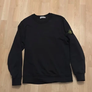 Svart tröja från Stone Island - Säljer en stilren svart tröja från Stone Island med deras ikoniska märke på ärmen. Tröjan har långa ärmar och en klassisk rund halsringning. Perfekt för en avslappnad och trendig look. Skriv vid eventuella frågor.