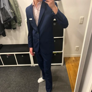 Mörkblå kostym - Snygg mörkblå kostym med kavaj och byxor. Kavajen har två knappar och en bröstficka med vit näsduk. Perfekt för studenten eller annat formellt tillfälle. Köpt för 2500 kr. 