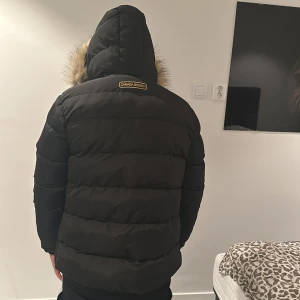 Svart dunjacka med päls från Canada Goose - Säljer en svart dunjacka från Canada Goose med pälsdetaljer på huvan. Jackan har en klassisk design med lång ärm och är perfekt för kalla dagar. Den har en dragkedja framtill och är märkt med Canada Goose-logotypen på ärmen.