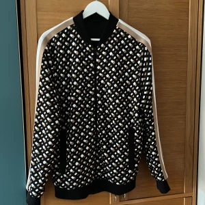 Hugo boss bomberjacka med dragkedja - Säljer en snygg Hugo Boss bomberjacka med ett unikt rutigt mönster i svart, beige och vitt. Jackan har långa ärmar och en dragkedja framtill. Den har ribbade muddar vid ärmslut och nederkant samt en vit rand längs ärmarna. Priset går att diskutera.