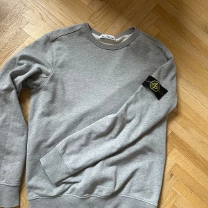Grå sweatshirt från Stone Island - Säljer en stilren grå sweatshirt från Stone Island med deras ikoniska märke på ärmen. Tröjan har en klassisk rund hals och långa ärmar, perfekt för en avslappnad look. Passar bra till både jeans och chinos.