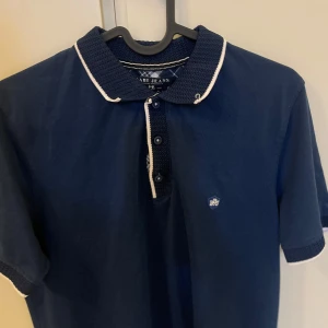 Mörkblå pikétröja från Rabi Jeans - Snygg mörkblå pikétröja från Rabi Jeans med vit kant på kragen och ärmsluten. Tröjan har en klassisk pikédesign med knappar framtill och en liten logotyp på bröstet. Perfekt för en stilren look.