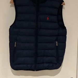 Mörkblå dunväst från Ralph Lauren - Snygg mörkblå dunväst från Ralph Lauren med dragkedja och broderad logga i rött på bröstet. Perfekt för kyligare dagar med sin quiltade design och praktiska fickor med dragkedja.
