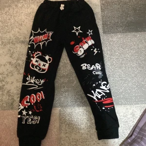 Svarta mjukisbyxor med grafiska tryck - Coola svarta mjukisbyxor med färgglada grafiska tryck och text som 'WOW', 'COOL' och 'KING'. Byxorna har elastisk midja med snörning och muddar vid bensluten. Perfekta för en avslappnad stil med en touch av attityd.