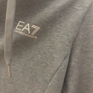 Grå huvtröja från Emporio Armani - Säljer en stilren grå huvtröja från Emporio Armani med dragkedja och justerbar huva. Tröjan har en diskret logga på bröstet och en snygg dragkedja med EA7-detalj. 