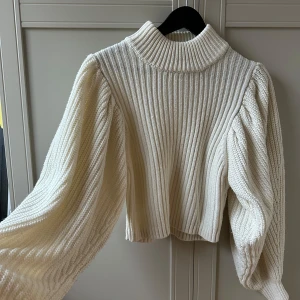 Vit ribbad stickad tröja - Säljer en stilren vit stickad tröja med ribbad design och puffiga ärmar. Tröjan har en hög krage och är croppad, vilket ger en modern look. 