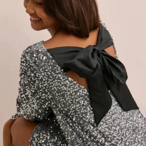 Glittrig silverklänning med svart rosett - Snygg glittrig klänning i silver med en stor svart rosett i ryggen. Klänningen har långa ärmar och en öppen rygg. Helt ny, med prislappen kvar💕☺️