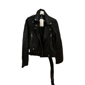 Svart skinnjacka från Bolongaro Trevor  - Snygg svart skinnjacka från Bolongaro med coola detaljer som dragkedjor och knappar. Jackan har en klassisk bikerstil med bälte i midjan och långa ärmar. Perfekt för att ge din outfit en edgy touch.