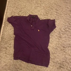 Lila pikétröja från U.S. Polo Assn. - Säljer en snygg lila pikétröja från U.S. Polo Assn. i storlek L. Tröjan är tillverkad i 100% bomull och har en klassisk krage med knappar. Perfekt för en avslappnad stil.