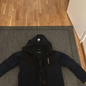 Mörkblå jacka från Sail Antarctica - Säljer en mörkblå jacka från Sail Antarctica med huva och dragkedja. Perfekt för kyliga dagar med sin pufferdesign. Jackan har en expeditionstema med en cool patch på ärmen. Pris kan diskuteras 