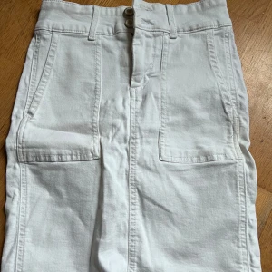 Vit jeanskjol med fickor - Snygg vit jeanskjol med två stora framfickor och knappstängning. Kjolen har en klassisk design med en rak passform och en diskret logotyp på baksidan. Perfekt för en stilren look.