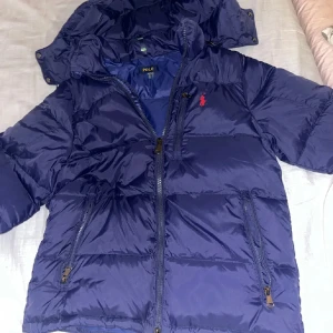 Blå dunjacka från Polo  - Säljer en blå dunjacka från Polo med avtagbar huva och dragkedja. Jackan har en broderad logga på bröstet och praktiska fickor med dragkedja. Perfekt för kyliga dagar.