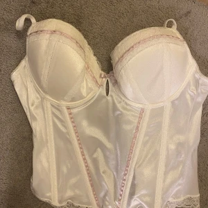 Vit korsett med spetsdetaljer - Elegant vit korsett med spetsdetaljer och vadderade kupor. Den har justerbara axelband och en fin glansig yta. Perfekt som fest topp
