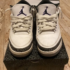 Nike Air Jordans 3 - Säljer ett par klassiska Nike Air Jordans i vitt med lila detaljer. Skorna har snörning och en unik mönstrad design på sidorna. Perfekta för den som vill ha en stilren och sportig look.