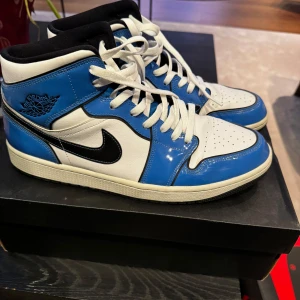 Nike Air Jordan 1 Mid SE i blå och vit - Snygga Nike Air Jordan 1 Mid SE sneakers i blått och vitt med svarta detaljer. Skorna har en klassisk snörning och är tillverkade i skinn. Knappt använda. 