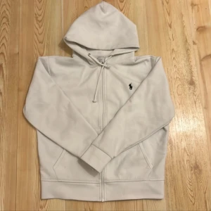 Vit hoodie från Ralph Lauren - Säljer en stilren vit hoodie från Ralph Lauren med dragkedja och klassisk logga på bröstet. Perfekt för en avslappnad look. Den har långa ärmar och en praktisk huva med dragsko.