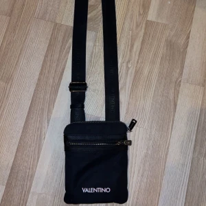 Svart axelväska från Valentino - Snygg svart axelväska från Valentino med justerbar rem och dragkedja. Finns 3 olika fickor med dragkedja på utsidan, 2 fram till och 1 på baksidan. Finns ytterligare ett innerfack inne i väskan