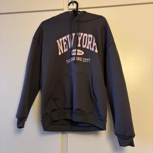 Mörkgrå hoodie med New York-tryck - Säljer en mörkgrå hoodie med texten 'New York' i vitt och rosa på framsidan. Hoodien har en stor ficka framtill och långa ärmar. Perfekt för en avslappnad stil.