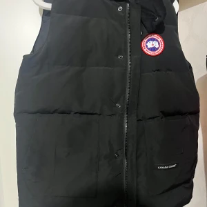 Svart dunväst från Canada Goose - Säljer en svart dunväst från Canada Goose med dragkedja och det klassiska märket på bröstet. Västen är perfekt för kyligare väder och har en stilren design.