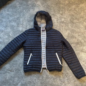 Mörkblå pufferjacka från Colmar - Säljer en snygg mörkblå pufferjacka från Colmar med ljusgrå insida. Jackan har en huva och dragkedja framtill samt två praktiska fickor med dragkedjor. Perfekt för kyliga dagar!