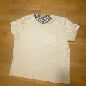 Vit t-shirt med blommigt mönster - Säljer en vit t-shirt med en blommig detalj runt halsen. T-shirten har en bröstficka och korta ärmar. Perfekt för en avslappnad stil.