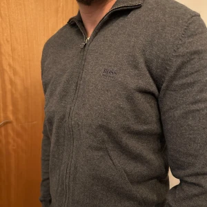 Grå kofta från Hugo Boss - Snygg grå kofta från Hugo Boss med dragkedja framtill. Perfekt för en stilren look. Den har långa ärmar och en diskret logga på bröstet. Passar bra till både vardag och mer uppklädda tillfällen.