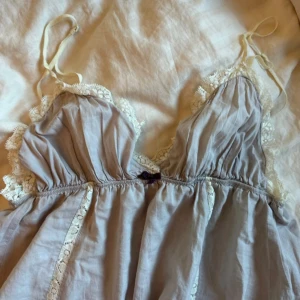 Ljuslila/grå klänning med spetsdetaljer - Charmig ljusgrå klänning med spetsdetaljer och broderi på baksidan. Klänningen har smala axelband och en luftig passform. Perfekt för en avslappnad och romantisk look.