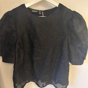 Svart blus från Pieces - Snygg svart blus från Pieces med korta puffärmar och ett diskret blommigt mönster. Perfekt för att ge en elegant touch till din outfit. Blusen har en rund halsringning och är tillverkad i ett lätt material.