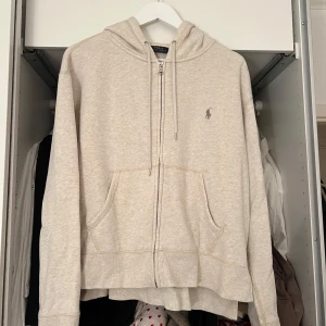 Beige hoodie från Polo Ralph Lauren - Säljer en stilren beige hoodie från Polo Ralph Lauren. Den har en dragkedja framtill och en klassisk logga på bröstet. Perfekt för en avslappnad look med sina långa ärmar och bekväma passform. Passar S-L