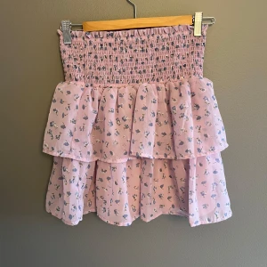 Rosa blommig kjol med volang - Charmig rosa kjol med blommigt mönster och volangdetaljer. Den har en smockad midja för en bekväm passform. Perfekt för en söt och romantisk stil 💗